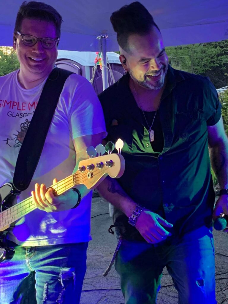 man in white t-shirt playing bass guitar and another man standing next to him smiling and laughing holding a microphone