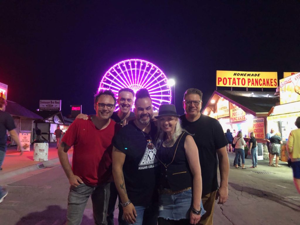 band members standing at a fair with a pink lit up ferris wheel behind them.
