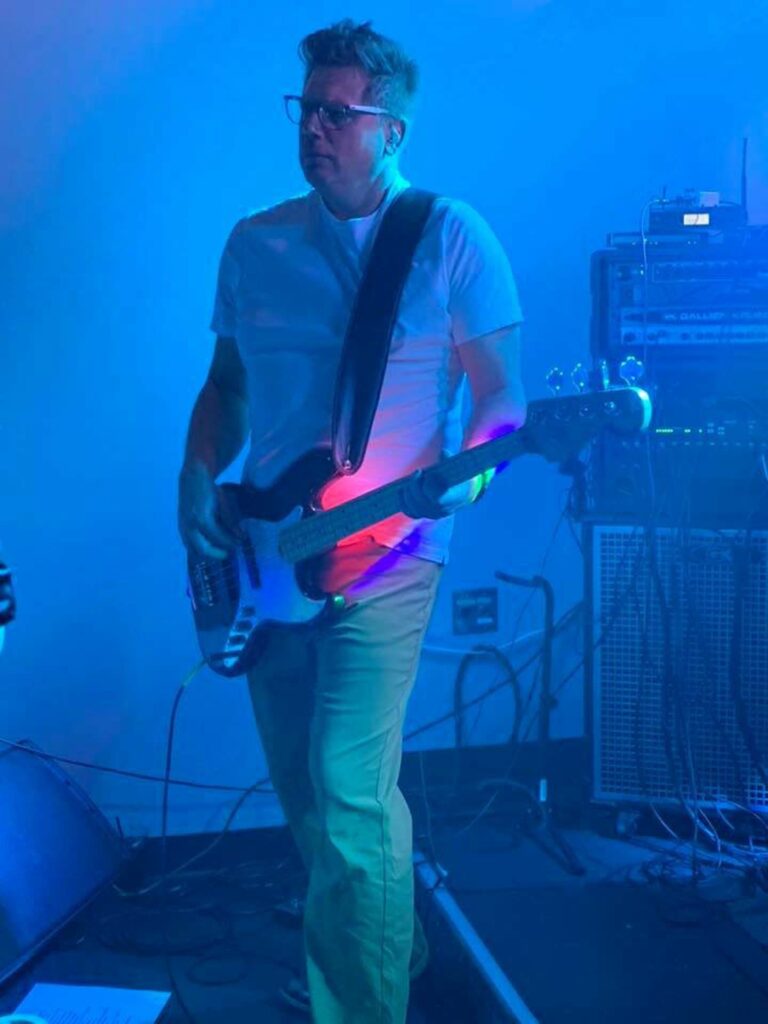 blue tone image of guy in a white t-shirt and tan colored jeans playing a bass guitar player