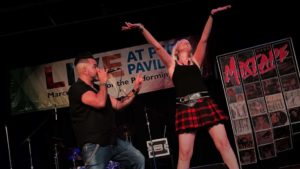 guy in black button t-shirt and jeans singing with girl in black tank and red and black checkered skirt standing to the right of him with her hands up in the air